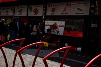 A group of people stand outside a store with signage in different languages and pictures of meat products. The store seems to specialize in various meats as indicated by the signs with illustrations. The entrance of the store is visible with some people interacting near it, possibly shopping or waiting.
