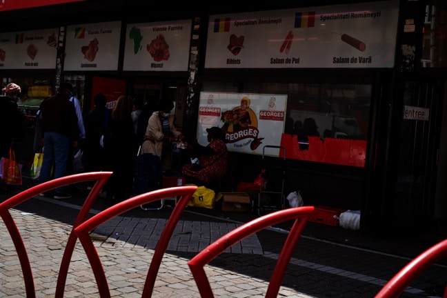 A group of people stand outside a store with signage in different languages and pictures of meat products. The store seems to specialize in various meats as indicated by the signs with illustrations. The entrance of the store is visible with some people interacting near it, possibly shopping or waiting.