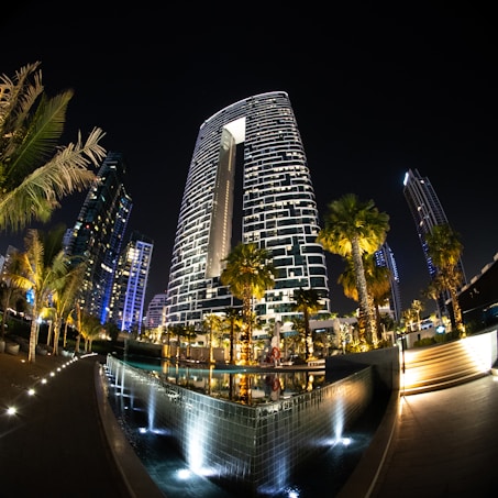 A futuristic cityscape at night featuring a prominent, modern skyscraper with a unique, illuminated facade. Surrounding the skyscraper are several other tall buildings, all glowing with various shades of light against the dark sky. In the foreground, palm trees and an elegantly lit water feature add to the opulent atmosphere.