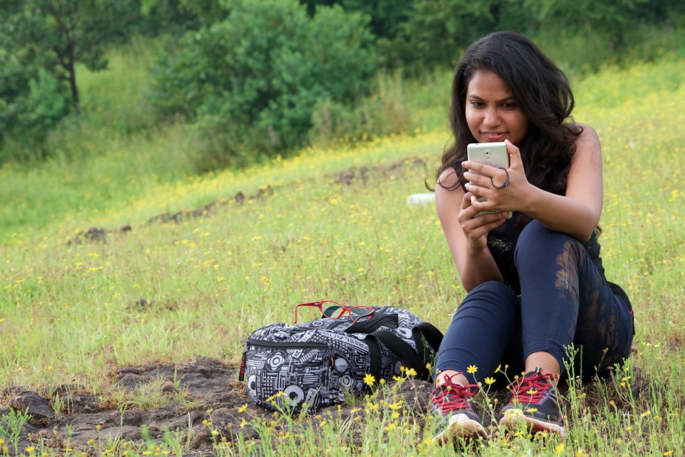 woman in black shirt and black pants sitting on grass field using smartphone