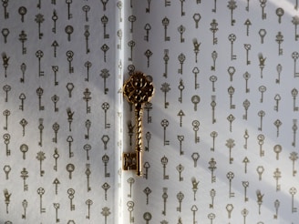A decorative pattern of vintage-style keys is printed on a light-colored surface. One prominent golden key is positioned in the center, casting a shadow on the surface.