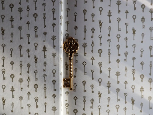 A decorative pattern of vintage-style keys is printed on a light-colored surface. One prominent golden key is positioned in the center, casting a shadow on the surface.