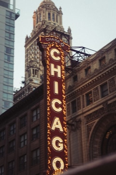 A historic theater sign illuminates with bright lights against the backdrop of a classic architectural building in an urban city setting. The tall tower in the background adds to the cityscape's grandeur, evoking a sense of nostalgia.