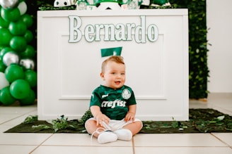 A baby dressed in a green football-themed outfit is sitting on the floor in front of a decorated table. The name 'Bernardo' is prominently displayed on the table. Green and white balloons are part of the festive decorations. The overall setting suggests a birthday or themed event.