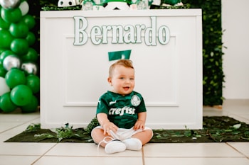 A baby dressed in a green football-themed outfit is sitting on the floor in front of a decorated table. The name 'Bernardo' is prominently displayed on the table. Green and white balloons are part of the festive decorations. The overall setting suggests a birthday or themed event.