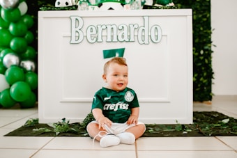 A baby dressed in a green football-themed outfit is sitting on the floor in front of a decorated table. The name 'Bernardo' is prominently displayed on the table. Green and white balloons are part of the festive decorations. The overall setting suggests a birthday or themed event.