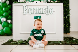 A baby dressed in a green football-themed outfit is sitting on the floor in front of a decorated table. The name 'Bernardo' is prominently displayed on the table. Green and white balloons are part of the festive decorations. The overall setting suggests a birthday or themed event.