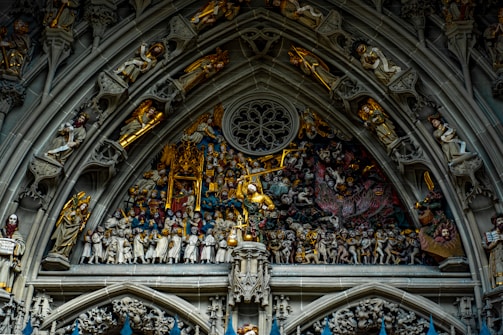 An intricately detailed relief depicting a religious scene, featuring numerous figures and ornate carvings. The architectural structure includes arches adorned with figures on both sides and a central scene filled with characters, some with golden accents, in a dramatic tableau.