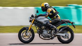 A motorcyclist wearing a helmet and protective gear rides a Ducati motorcycle on a racetrack. The motion blur in the background suggests high speed. The rider is dressed in a yellow, black, and blue outfit, and the motorcycle has a sleek, modern design.