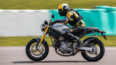 A motorcyclist wearing a helmet and protective gear rides a Ducati motorcycle on a racetrack. The motion blur in the background suggests high speed. The rider is dressed in a yellow, black, and blue outfit, and the motorcycle has a sleek, modern design.