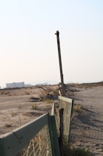 An old fence runs along a barren, sandy landscape. The fence is made of wire mesh attached to bent and rusted metal posts. Sparse vegetation and dry grass cover parts of the sand, while an urban area with buildings is visible in the hazy background. The scene suggests abandonment and decay.