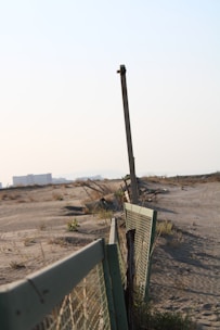 An old fence runs along a barren, sandy landscape. The fence is made of wire mesh attached to bent and rusted metal posts. Sparse vegetation and dry grass cover parts of the sand, while an urban area with buildings is visible in the hazy background. The scene suggests abandonment and decay.