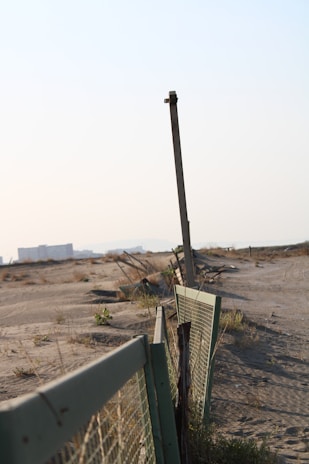 An old fence runs along a barren, sandy landscape. The fence is made of wire mesh attached to bent and rusted metal posts. Sparse vegetation and dry grass cover parts of the sand, while an urban area with buildings is visible in the hazy background. The scene suggests abandonment and decay.