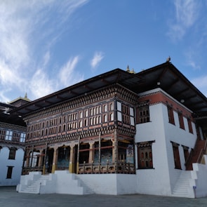 A traditional Bhutanese building adorned with intricate woodwork and colorful decorations. The structure features ornate patterns and architectural details typical of Bhutanese architecture. It stands against a backdrop of a clear blue sky with wispy clouds.