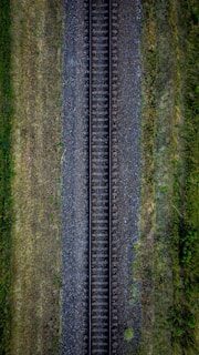 An aerial view of a single railway track running straight through a landscape surrounded by greenery and grass. The track is flanked by gravel on both sides, and the vegetation appears lush and evenly distributed.