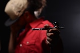 A person wearing a beige cap and a red jacket holds a metallic airbrush tool in focus, with a dark, blurred background.