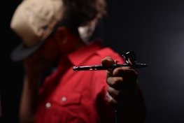 A person wearing a beige cap and a red jacket holds a metallic airbrush tool in focus, with a dark, blurred background.