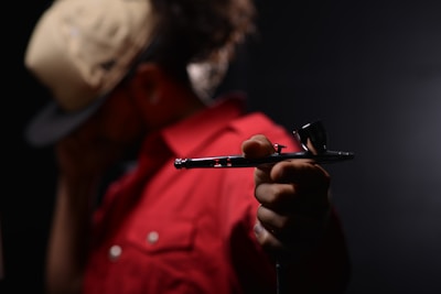 A person wearing a beige cap and a red jacket holds a metallic airbrush tool in focus, with a dark, blurred background.