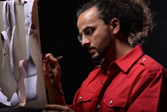 A person with glasses and a beard is using an airbrush to paint on a canvas. They are wearing a bright red shirt and have their hair tied back. The canvas has paper cutouts attached to it.
