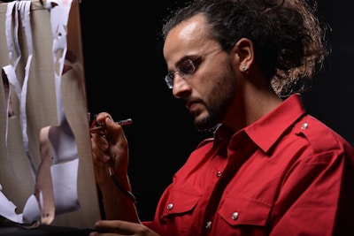 A person with glasses and a beard is using an airbrush to paint on a canvas. They are wearing a bright red shirt and have their hair tied back. The canvas has paper cutouts attached to it.