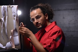 A person with glasses and a red shirt is holding an airbrush, engaged in a creative activity. The background is dark, and there is a bright light source to the side. Torn paper pieces hang in front of the person, creating an interesting visual effect.
