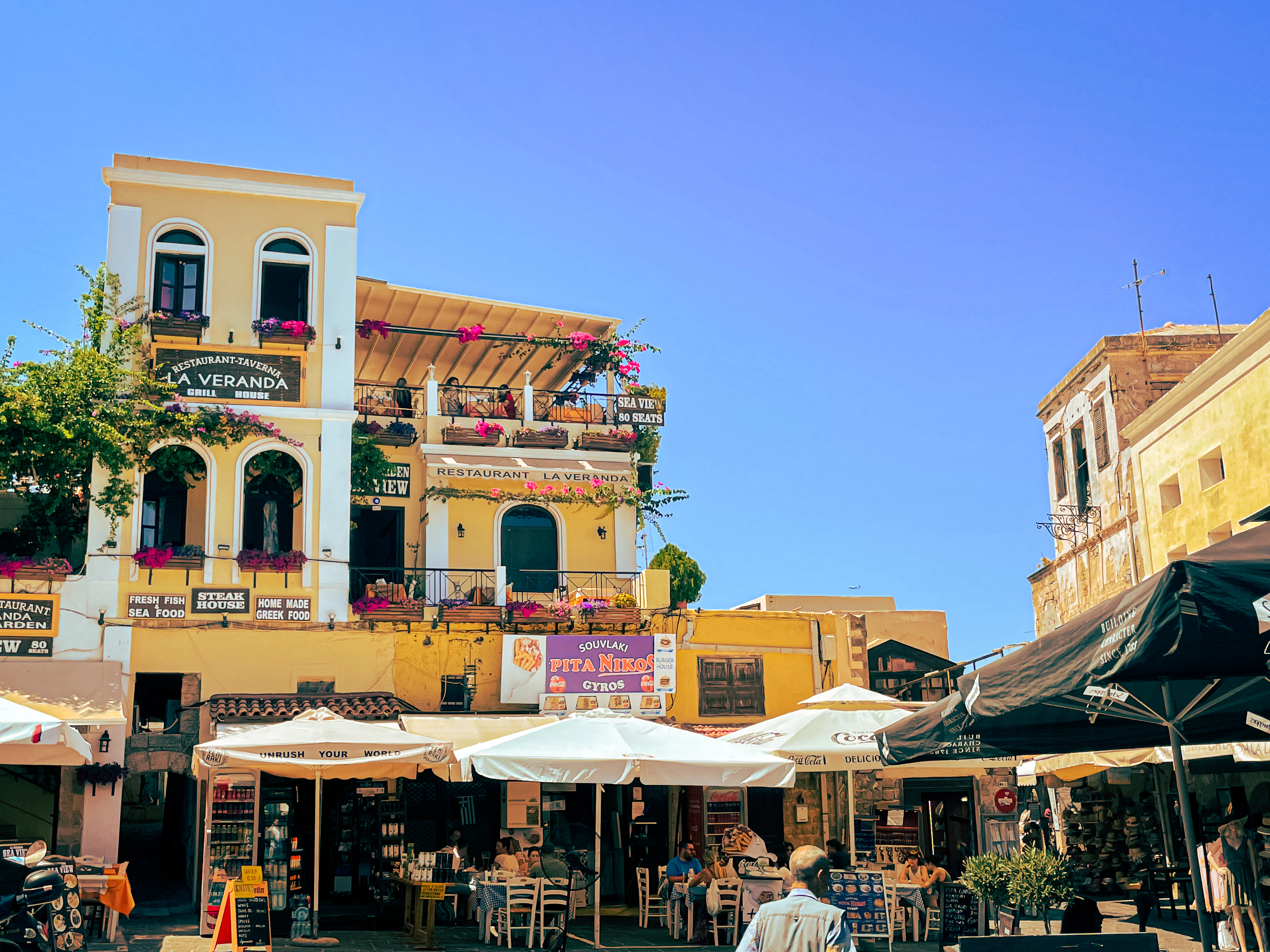 A square in Rhodes Old Town, ochre buildings and awnings above cafe tables set out on the flagstones