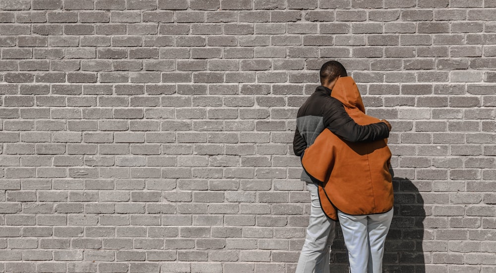 man in black and brown jacket and white pants standing beside brick wall