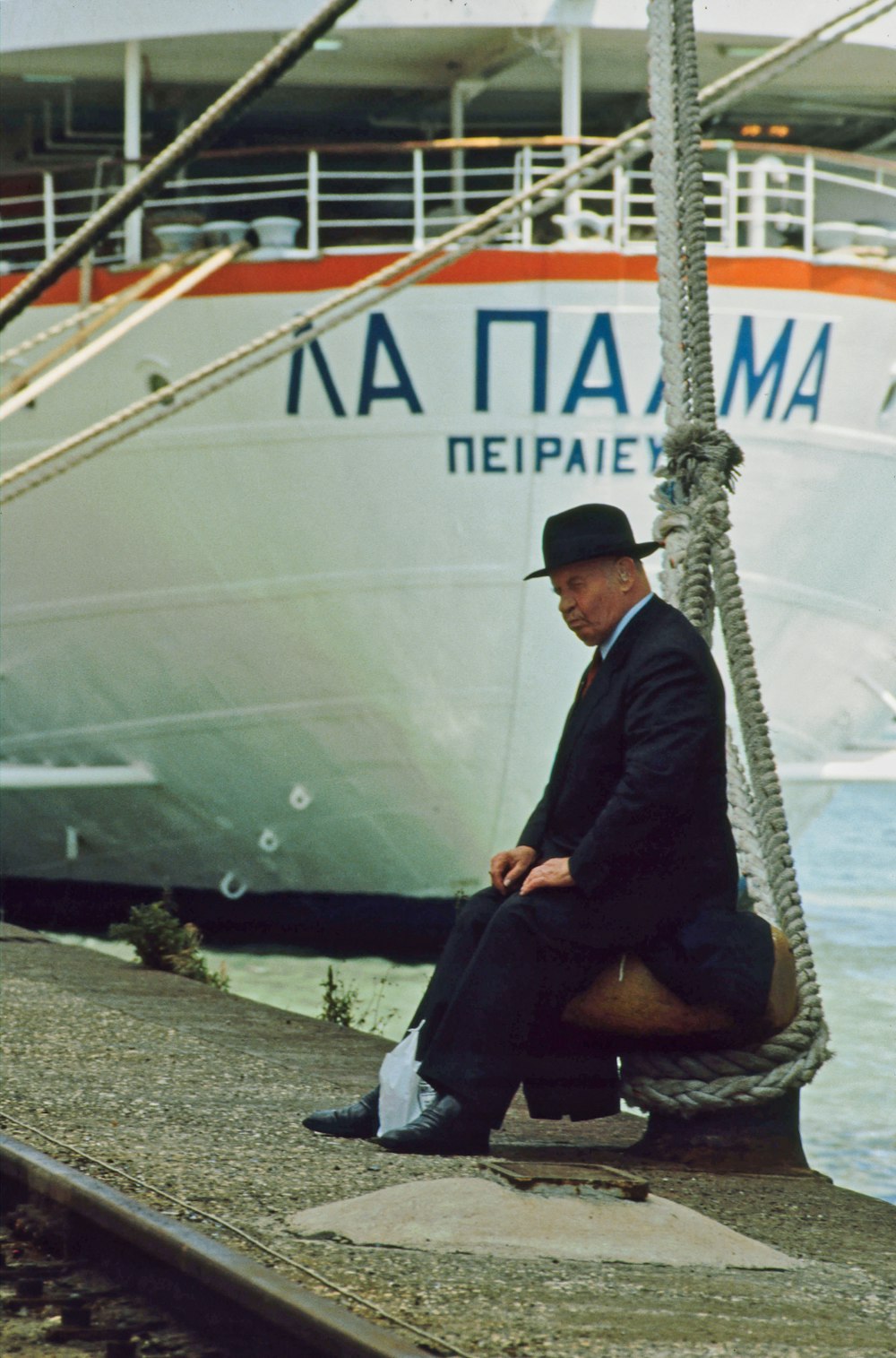 man in black jacket sitting on gray concrete pavement near white ship during daytime