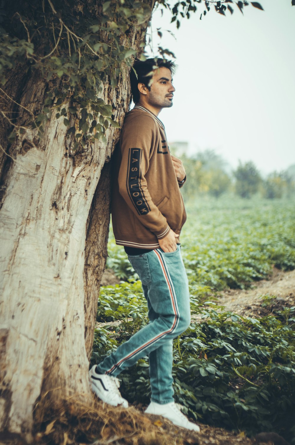 man in brown long sleeve shirt and blue denim jeans standing beside brown tree during daytime