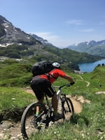 A cyclist on a mountain bike rides down a narrow trail surrounded by lush green hills and mountains. The scene overlooks a blue lake nestled among the mountains under a clear blue sky.