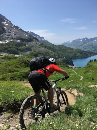 A cyclist on a mountain bike rides down a narrow trail surrounded by lush green hills and mountains. The scene overlooks a blue lake nestled among the mountains under a clear blue sky.