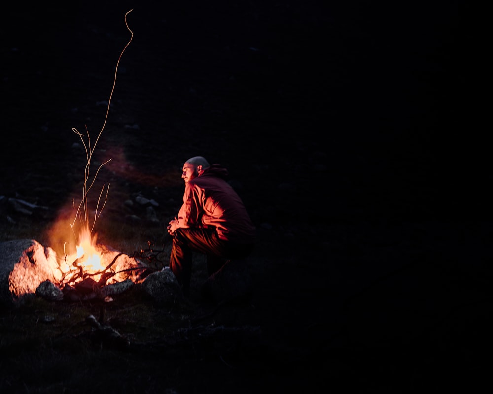 man in brown jacket sitting on ground with bonfire during night time