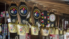 A collection of decorative cowbells hanging from a wooden structure, each adorned with intricate designs and the word 'Switzerland'. The bells feature images of Swiss landscapes, cows, and floral patterns, and are accompanied by leather straps with colorful embroidery.