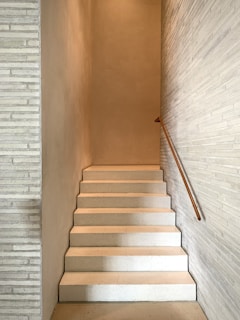 A minimalist indoor staircase with a smooth, light-colored finish, flanked by narrow walls. The design includes a simple wooden handrail on one side, creating a sense of simplicity and modern architectural style.