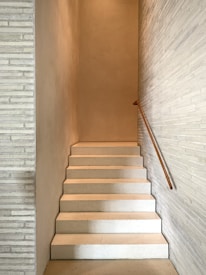 A minimalist indoor staircase with a smooth, light-colored finish, flanked by narrow walls. The design includes a simple wooden handrail on one side, creating a sense of simplicity and modern architectural style.