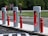 Several Tesla electric car charging stations are positioned in a row along a roadside. Bright red and white color designs accentuate the sleek, modern-looking chargers. Bright red bollards line the edge of the charging area, with signs indicating 'Tesla Vehicle Charging Only' and 'Do Not Enter' visible in the background. The area is surrounded by green grassy fields and trees, creating a contrast between the natural setting and the technological infrastructure.