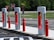 Several Tesla electric car charging stations are positioned in a row along a roadside. Bright red and white color designs accentuate the sleek, modern-looking chargers. Bright red bollards line the edge of the charging area, with signs indicating 'Tesla Vehicle Charging Only' and 'Do Not Enter' visible in the background. The area is surrounded by green grassy fields and trees, creating a contrast between the natural setting and the technological infrastructure.