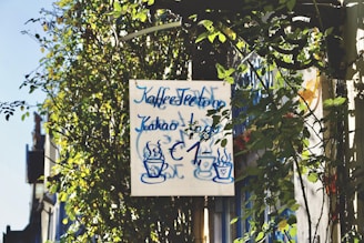A sign surrounded by lush green foliage displays handwritten text in blue, advertising coffee, tea, and cocoa for one euro. Two steaming cup illustrations are also present on the sign, which is set against a backdrop of climbing plants and a clear blue sky.