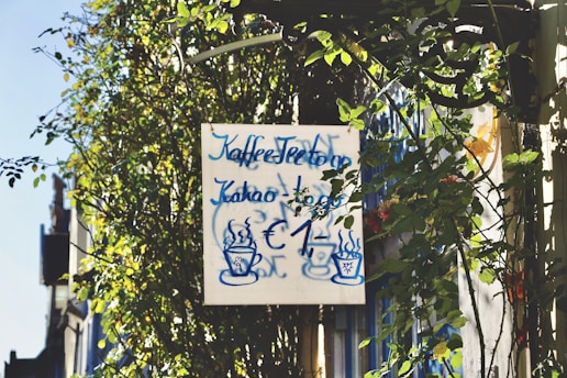 A sign surrounded by lush green foliage displays handwritten text in blue, advertising coffee, tea, and cocoa for one euro. Two steaming cup illustrations are also present on the sign, which is set against a backdrop of climbing plants and a clear blue sky.