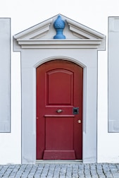 A red wooden door with a classic panel design is set within a white building with gray stone trim. Above the door is a decorative triangular pediment featuring a blue ornament. The door sits atop a cobblestone pathway.