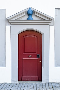 A red wooden door with a classic panel design is set within a white building with gray stone trim. Above the door is a decorative triangular pediment featuring a blue ornament. The door sits atop a cobblestone pathway.