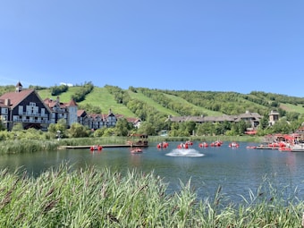 A scenic view of a lakeside resort with traditional-style buildings set against lush green hills. The lake is dotted with red paddle boats and has a small fountain near the center. Tall grasses border the foreground, with clear blue skies overhead.
