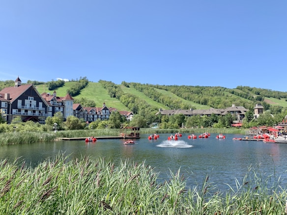 A scenic view of a lakeside resort with traditional-style buildings set against lush green hills. The lake is dotted with red paddle boats and has a small fountain near the center. Tall grasses border the foreground, with clear blue skies overhead.