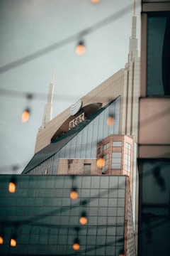 A modern skyscraper with sharp angles and a prominent logo near the top. The building features a glass facade reflecting the sky and surrounding structures. In the foreground, soft-focus lights create a bokeh effect, adding depth and atmospheric detail to the scene.