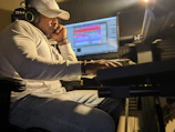 A person wearing a white shirt and a cap is intently focused on a computer screen with audio editing software. They are seated in a dimly lit room, wearing headphones, and appear to be engaged in music production.