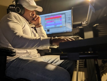 A person wearing a white shirt and a cap is intently focused on a computer screen with audio editing software. They are seated in a dimly lit room, wearing headphones, and appear to be engaged in music production.