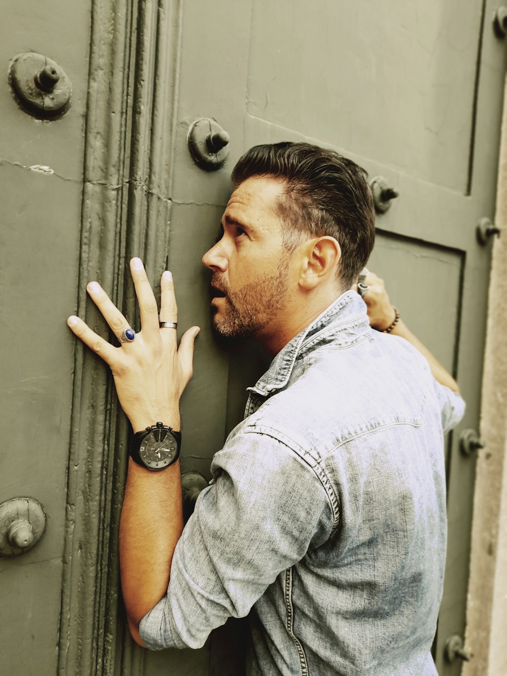 Hombre con camisa azul de mezclilla abotonada apoyada en una puerta de metal gris