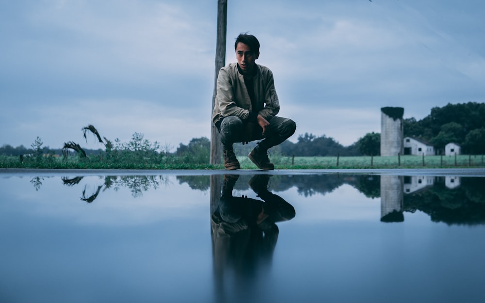 man in black jacket and black pants sitting on wooden post near body of water during