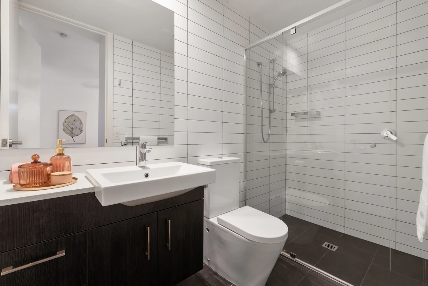 Compact modern bathroom with white horizontal wall tiles, a glass shower enclosure, wall-mounted shower fittings, white toilet and dark vanity.
