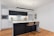 A modern kitchen with a minimalist design featuring white cabinetry and dark wood accents. The central island includes a white countertop and is complemented by two woven bar stools. Appliances like a microwave are integrated into the cabinetry, and decorative elements such as flowers and framed art are present. The lighting is contemporary, using a long, thin pendant light above the island.